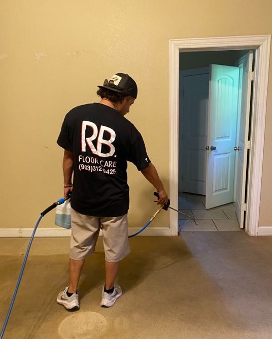Man in black shirt sprays carpet with a cleaning solution. He stands in front of an open doorway.