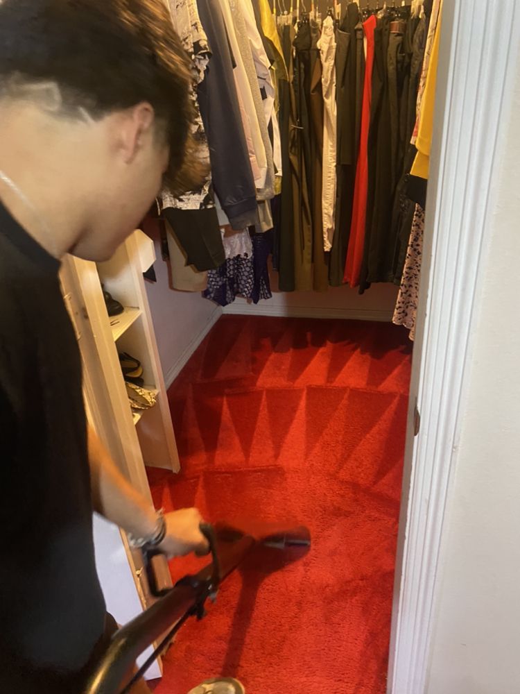 Person vacuuming a red carpeted closet; clothing hangs in the background.