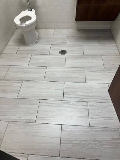 Bathroom with light gray wood-look tile floor, toilet, and drain. Dark wood-toned cabinet visible.