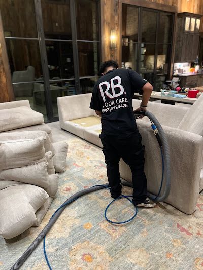 Man in black shirt vacuums a beige sofa on a patterned rug.