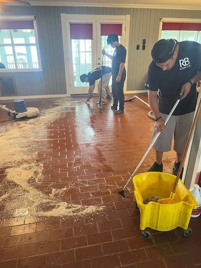 Several people cleaning a red brick floor with mops and buckets in a room with windows.