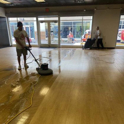 Two men cleaning a wooden floor with a floor buffer in a brightly lit room.