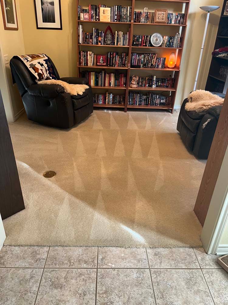 A beige carpeted room with two black recliners, a bookcase, and tile flooring. The carpet has a pattern.