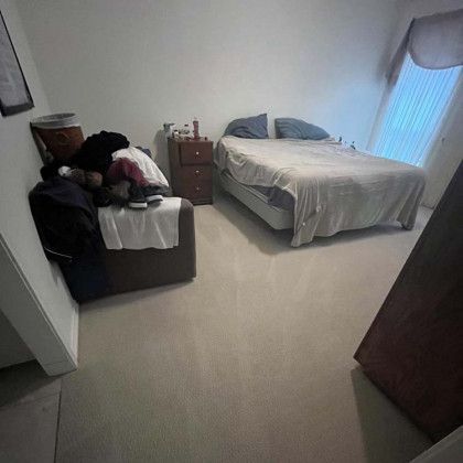 Bedroom with bed, nightstand, and a chair with clothes. Light-colored walls and carpet.