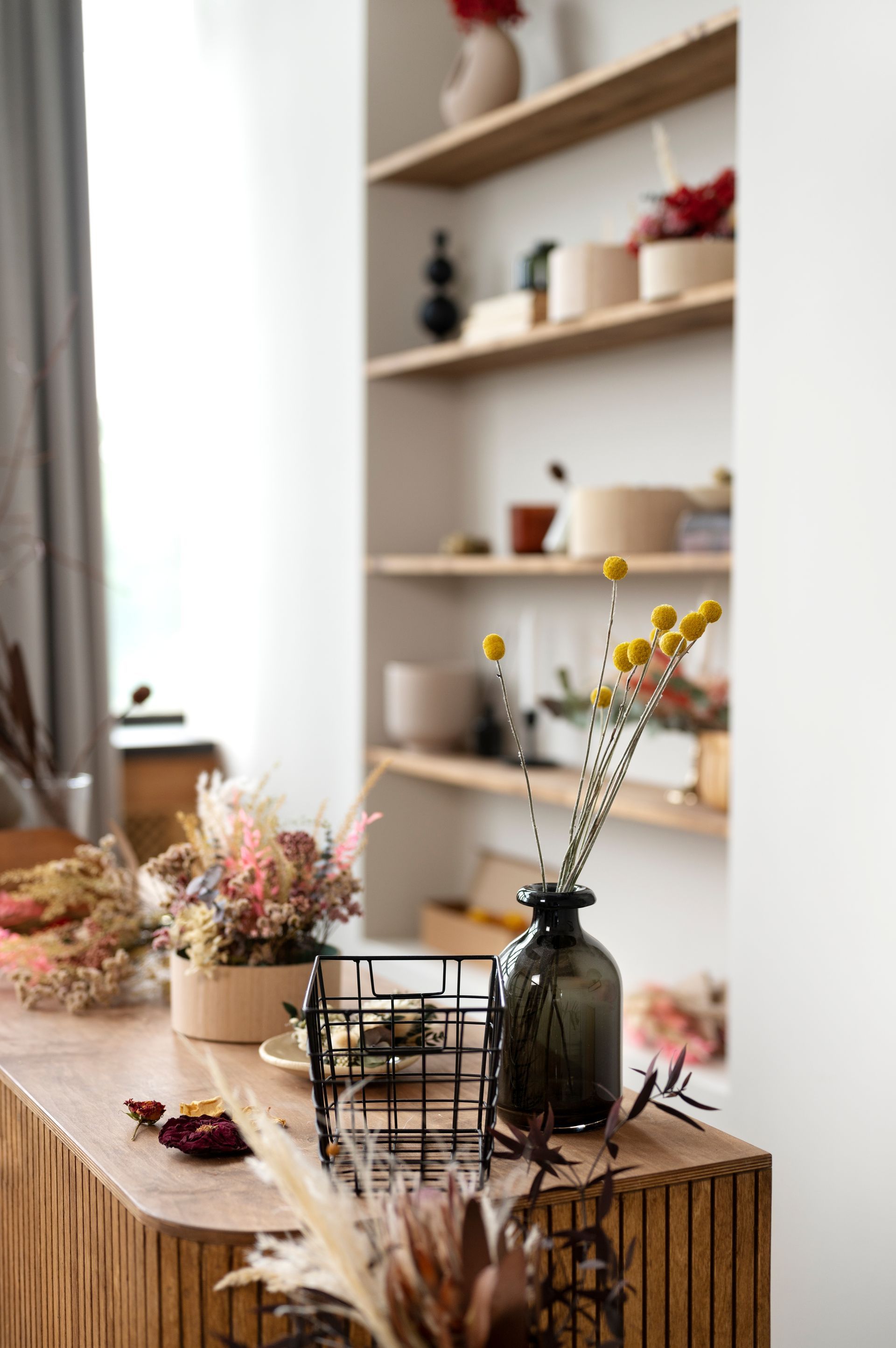 Mostrador de madera con jarrones de flores secas, una cesta de alambre y estantes al fondo.