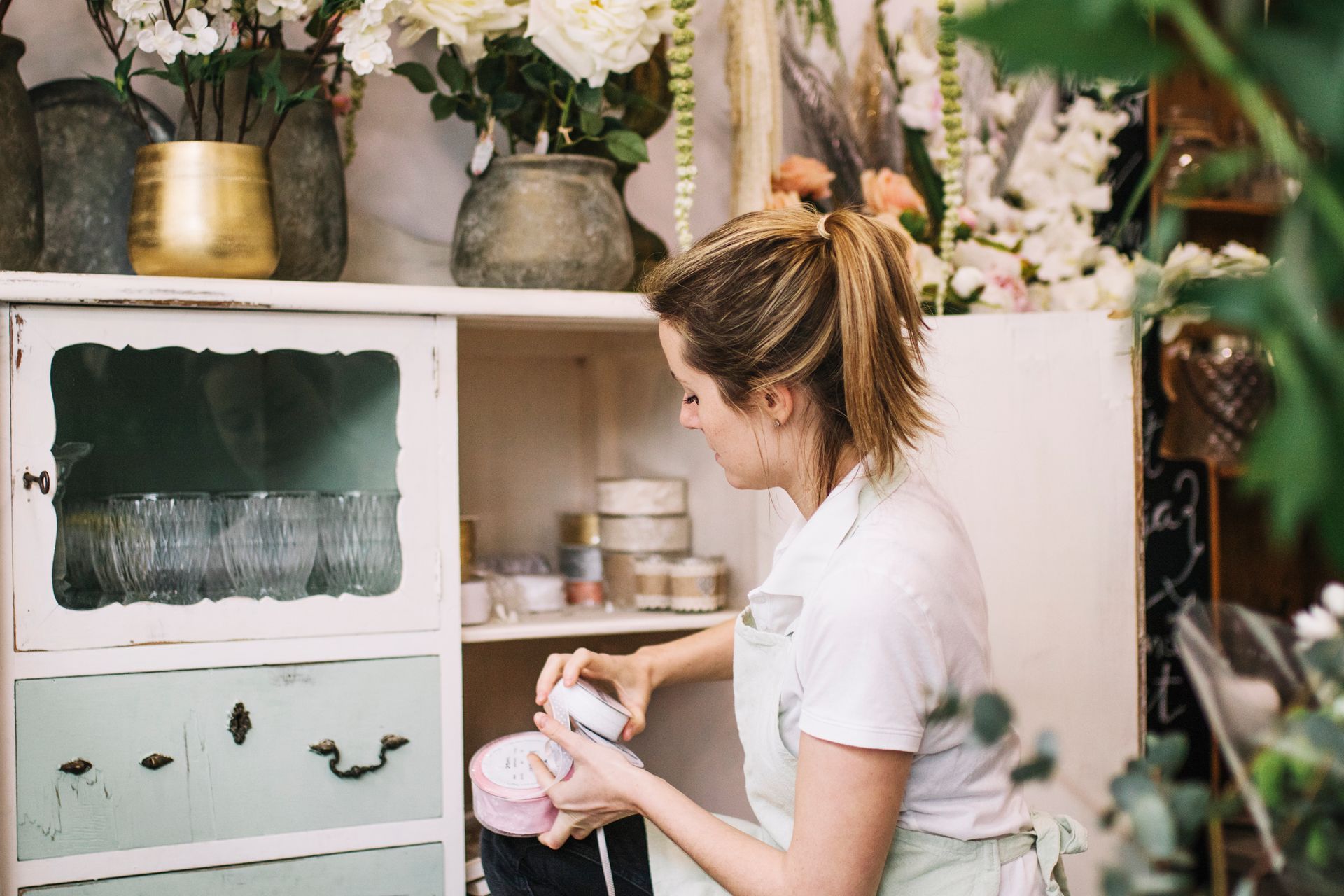 Mujer en una floristería, organizando suministros en un estante rodeado de flores.