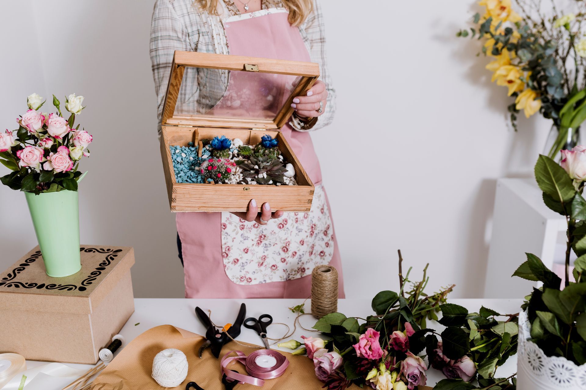 Mujer con delantal sosteniendo una caja de madera con flores secas, rodeada de flores y suministros sobre una mesa.