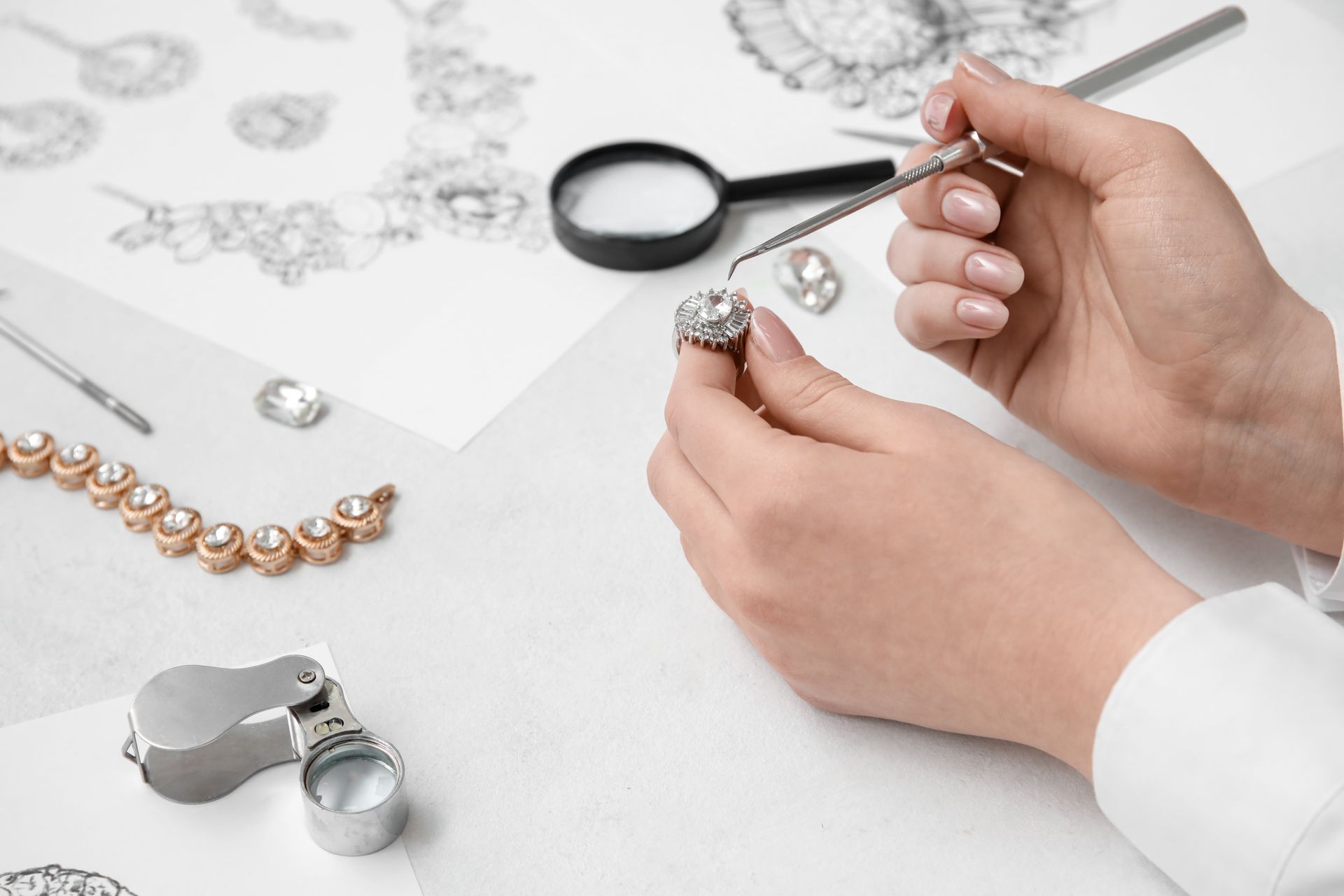 Hands using tweezers to inspect a diamond ring, jewelry design sketches, and tools on a white surface.