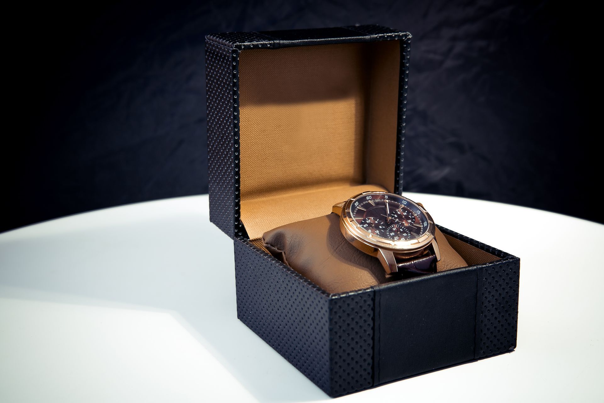 Black watch in a patterned box with a tan interior, on a white surface against a dark background.