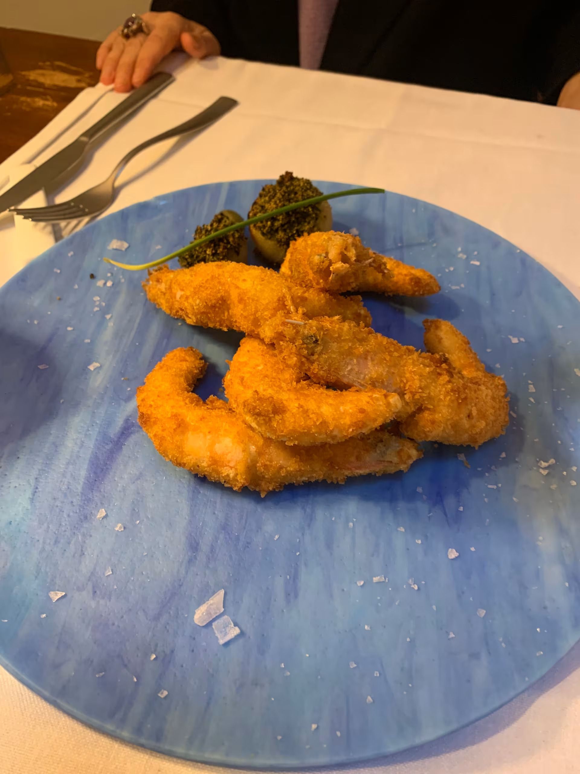 Gamberi fritti impanati su un piatto blu con guarnizione, su un tavolo con posate nelle vicinanze.