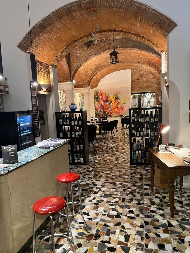 Un corridoio ad arco si apre su una caffetteria con tavoli, pavimento piastrellato e calde lampade a sospensione.