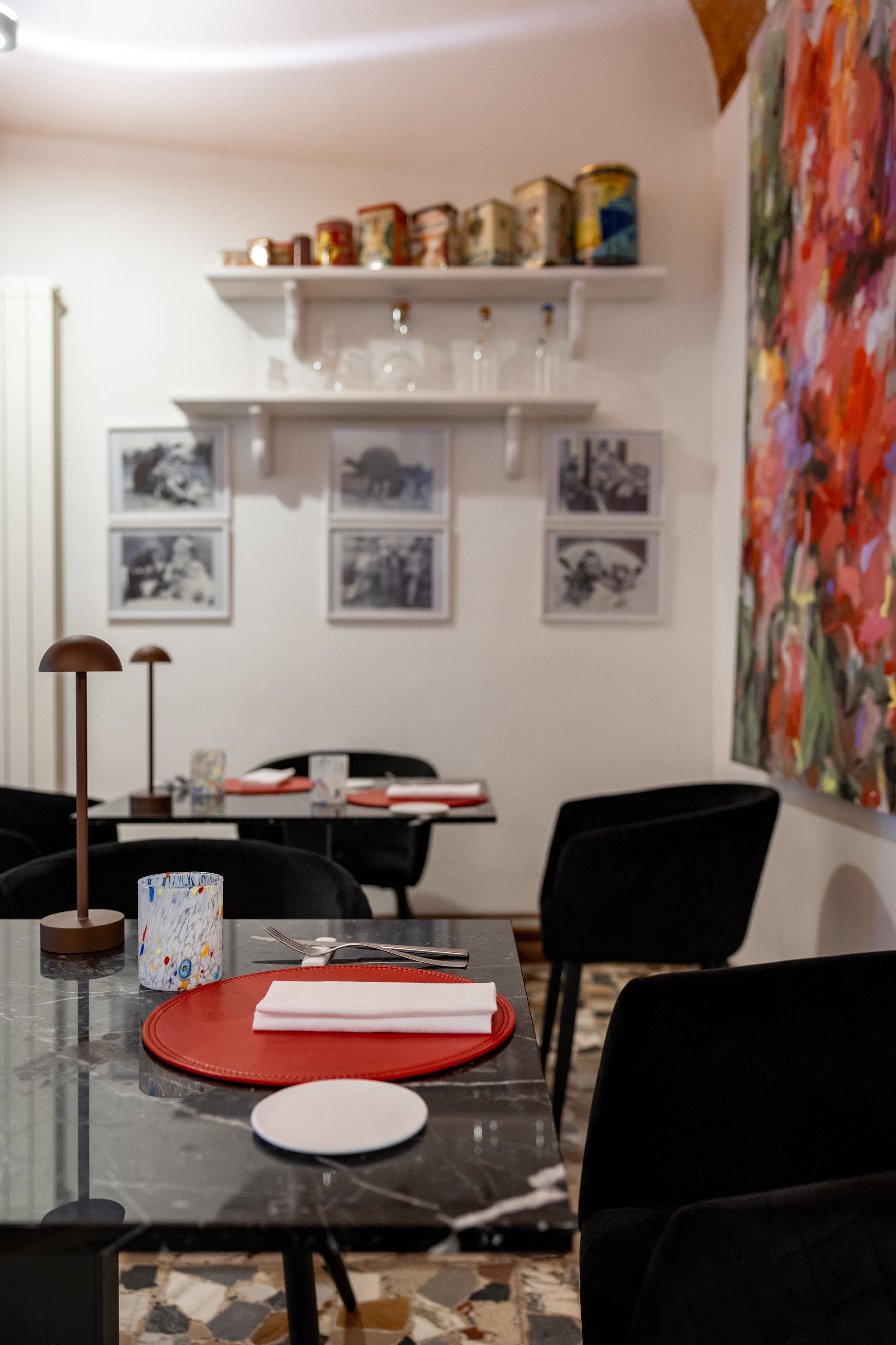 Accogliente angolo pranzo con sedie nere, tovagliette rosse, quadri incorniciati alle pareti, mensole e un dipinto colorato.