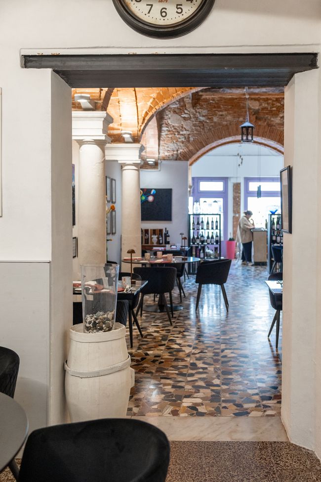 Un corridoio ad arco si apre su una caffetteria con tavoli, pavimento piastrellato e calde lampade a sospensione.