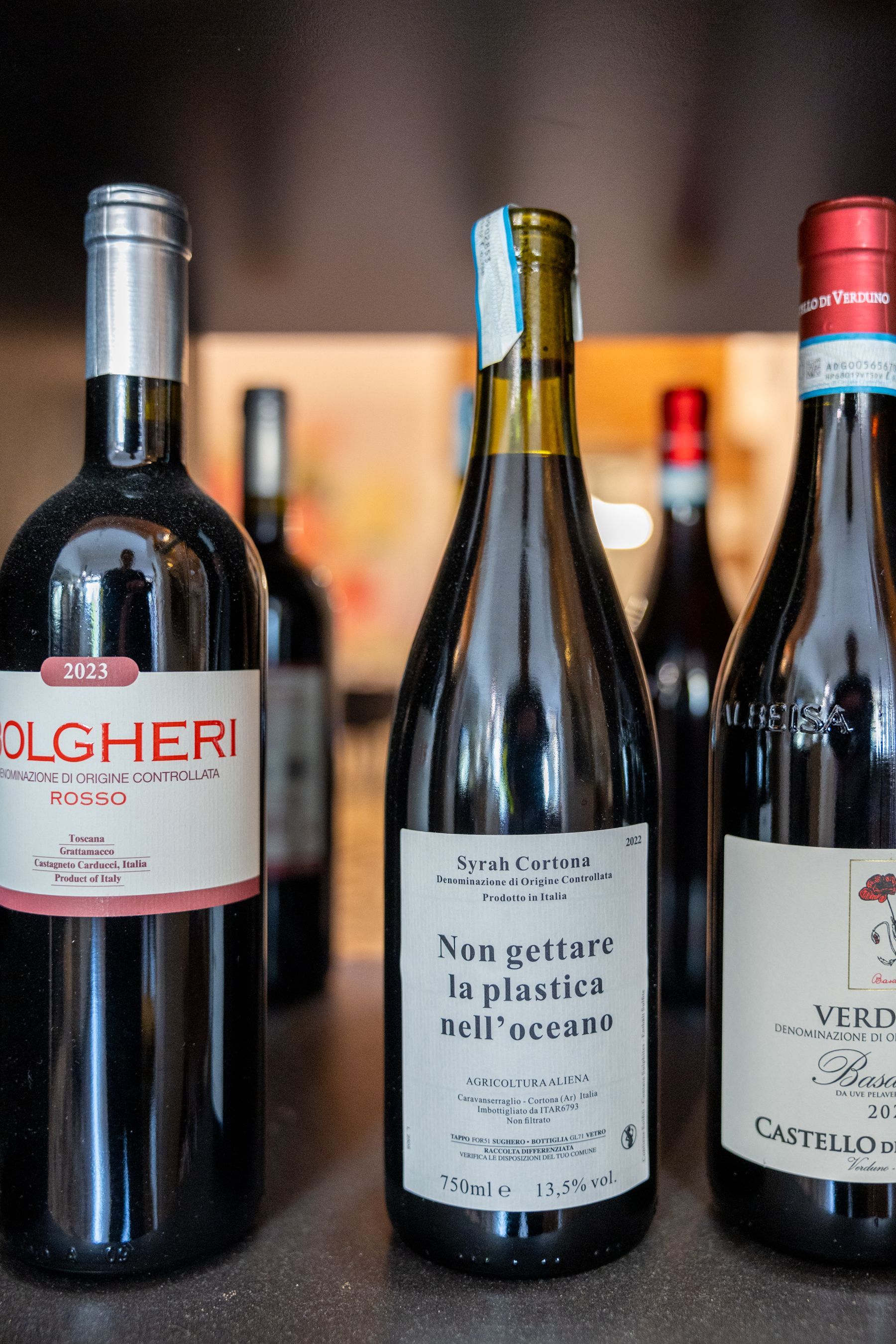 Tre bottiglie di vino esposte su uno scaffale, con una bottiglia rossa a sinistra e una bottiglia con tappo rosso a destra.