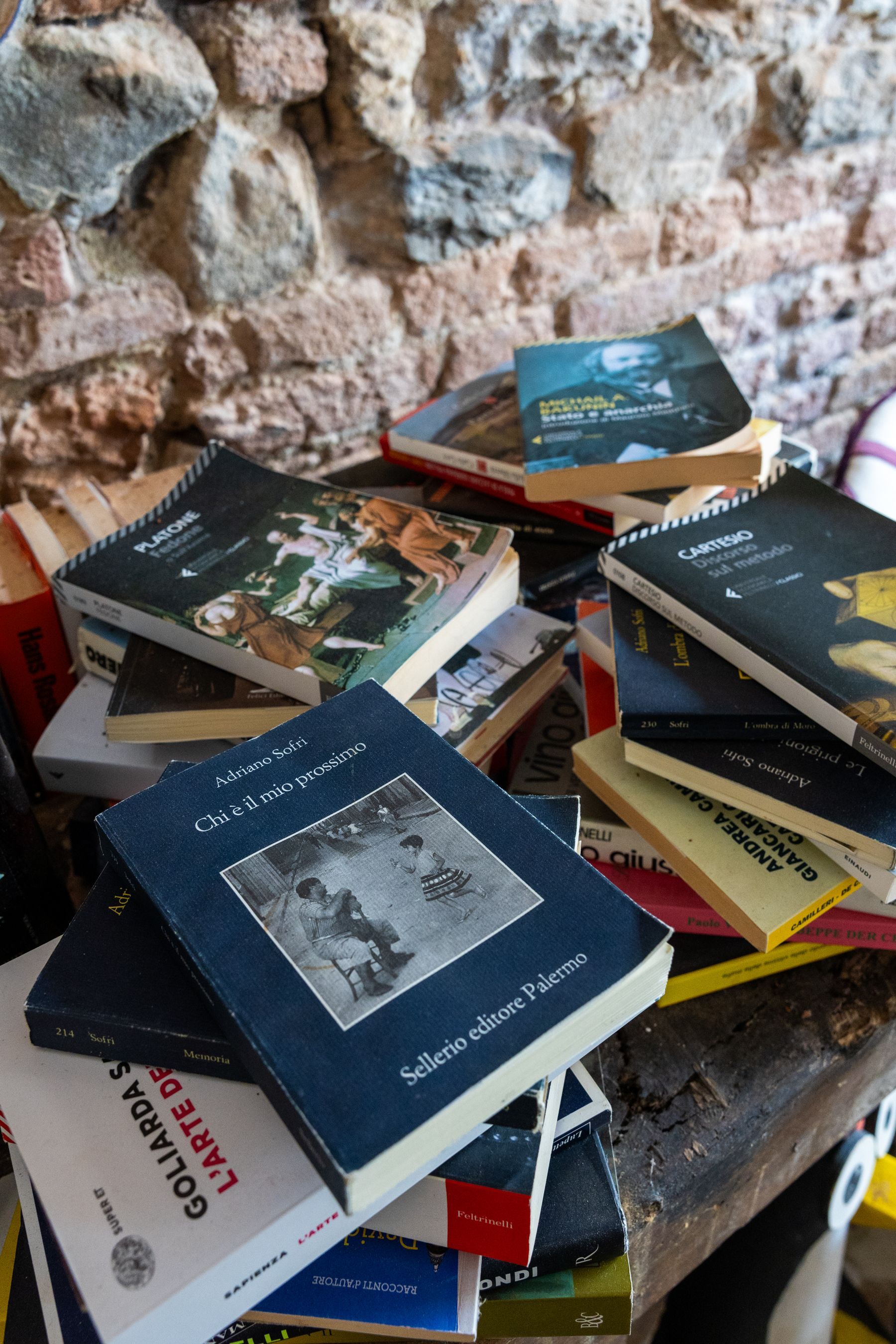Pila di libri di vario genere accatastati su una superficie di pietra addossata a un muro di pietra.