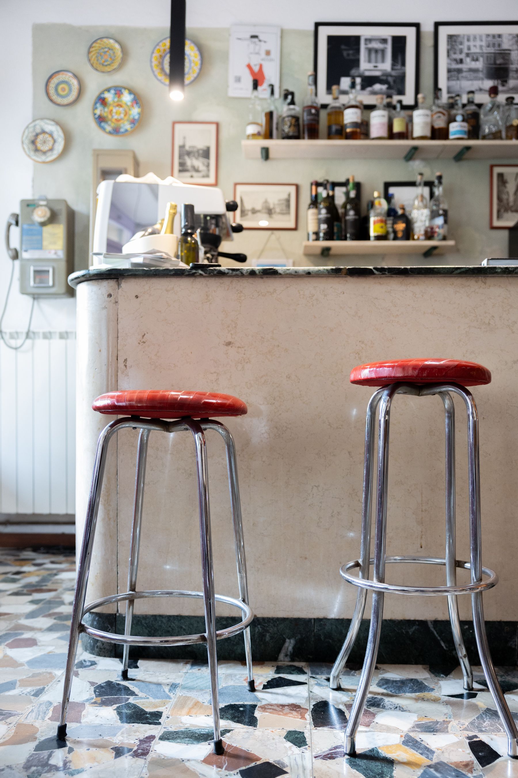 Due sgabelli rossi al bancone di un accogliente caffè, con scaffali pieni di bottiglie e foto incorniciate alle loro spalle.
