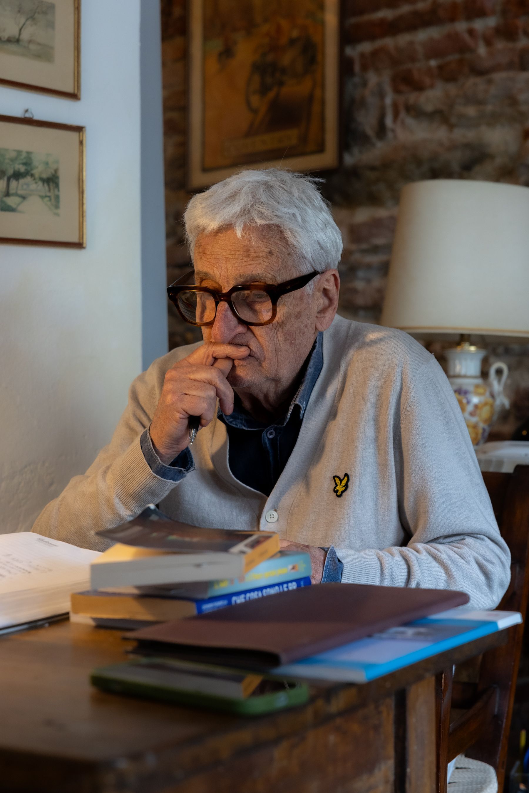 Un uomo anziano con gli occhiali legge a un tavolo di legno in una stanza accogliente, con libri e una lampada nelle vicinanze.
