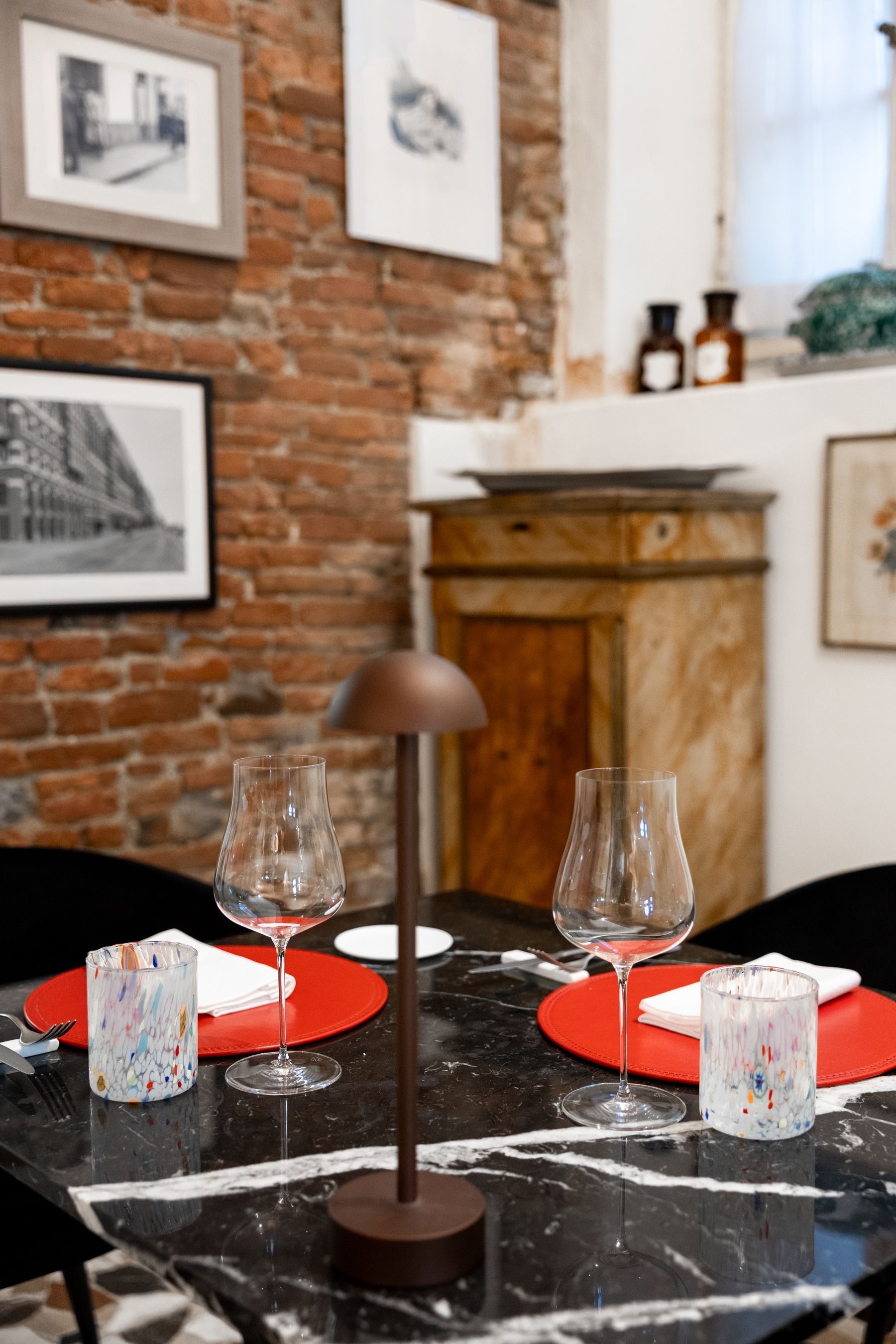 Accogliente tavolo da ristorante apparecchiato con tovagliette rosse, bicchieri da vino, candele e uno sfondo con muro di mattoni.
