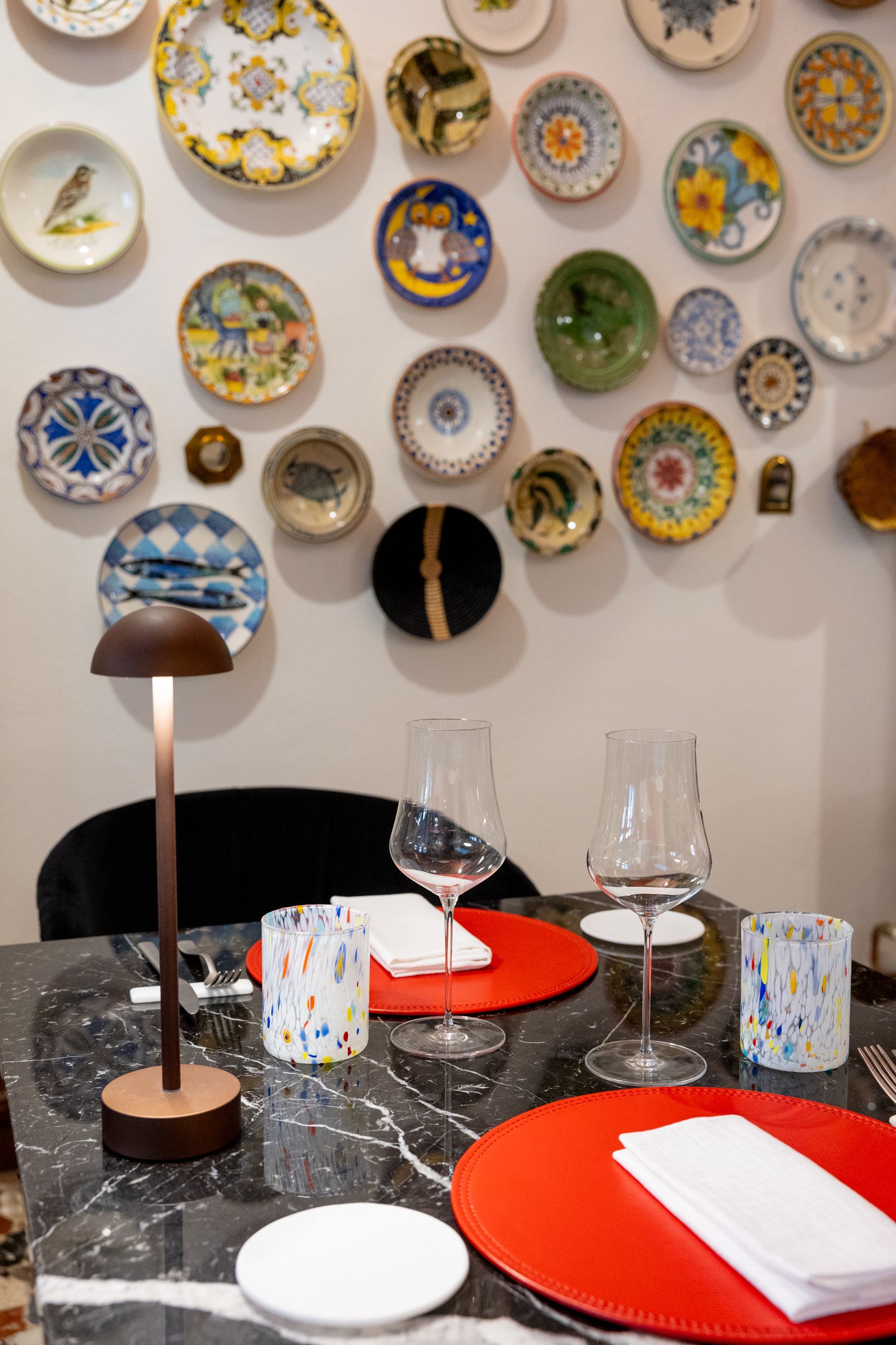 Tavolo da pranzo apparecchiato con tovagliette rosse e bicchieri sotto una parete di piatti decorativi.