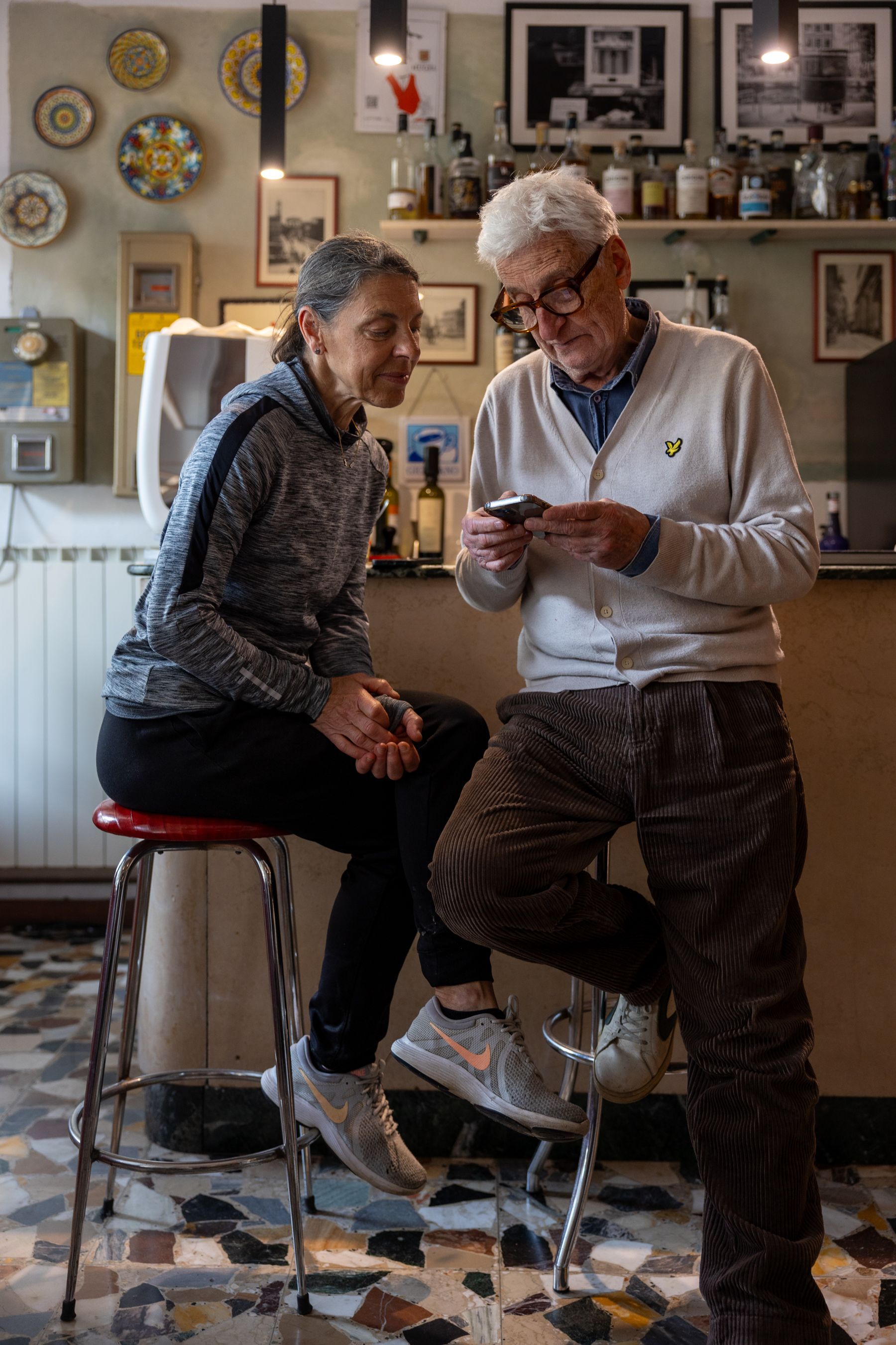 Due persone sorridenti in una cucina accogliente, sedute su degli sgabelli, guardano un telefono.