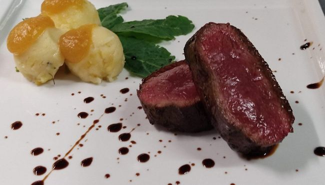 Fette di carne scottate servite con purè di patate, guarnizione verde e gocce di salsa su un piatto bianco.