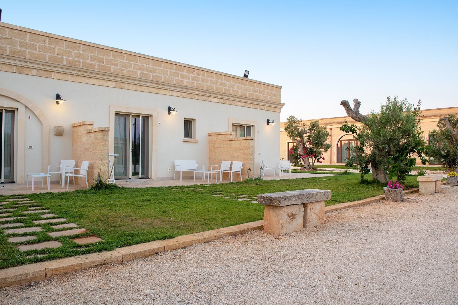 Vista esterna di un edificio bianco con un prato verde, sedie bianche e un ulivo in Puglia, Italia.