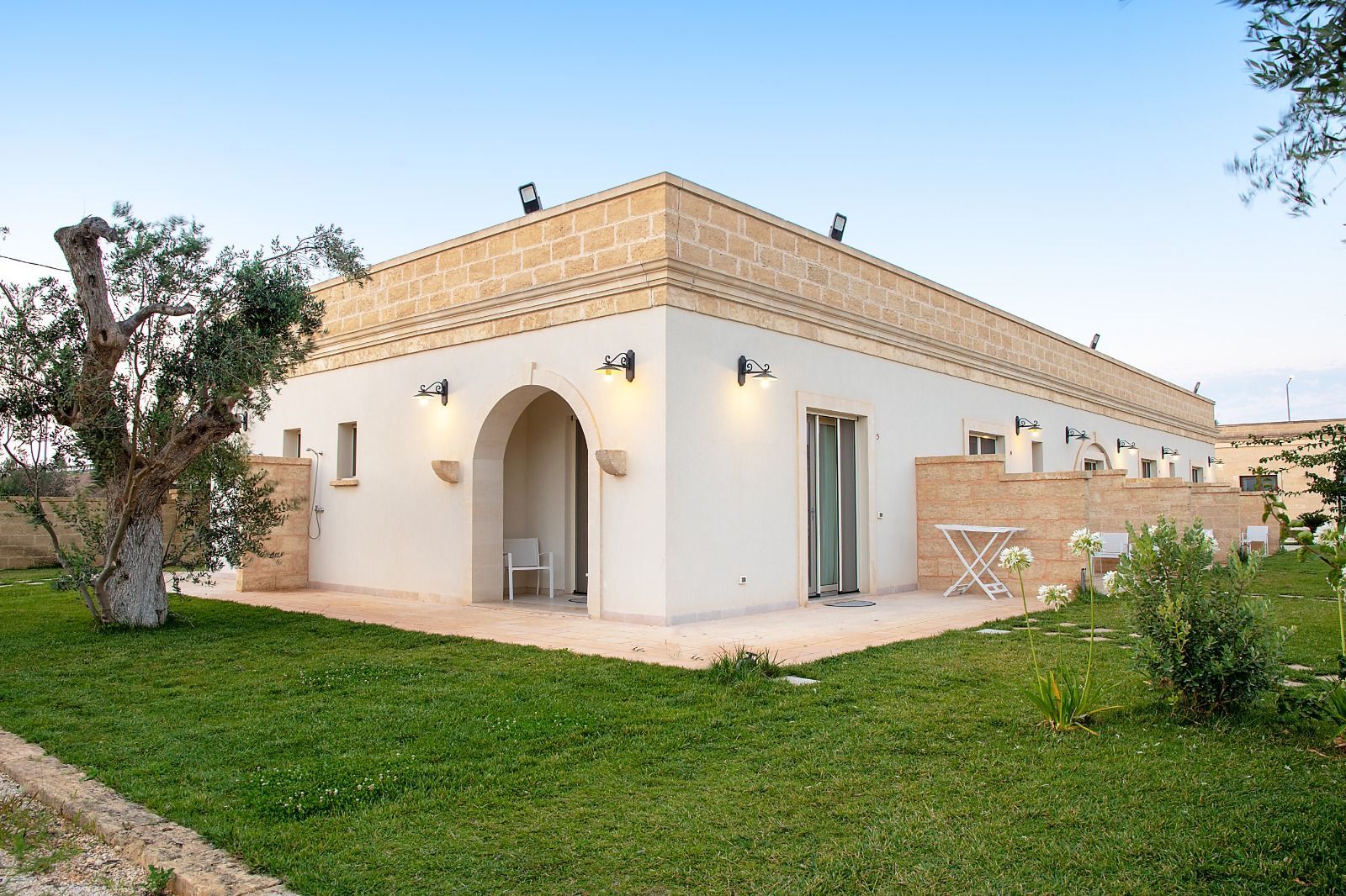Edificio con pareti bianche, ingresso ad arco, finiture in pietra, prato verde e cielo azzurro.