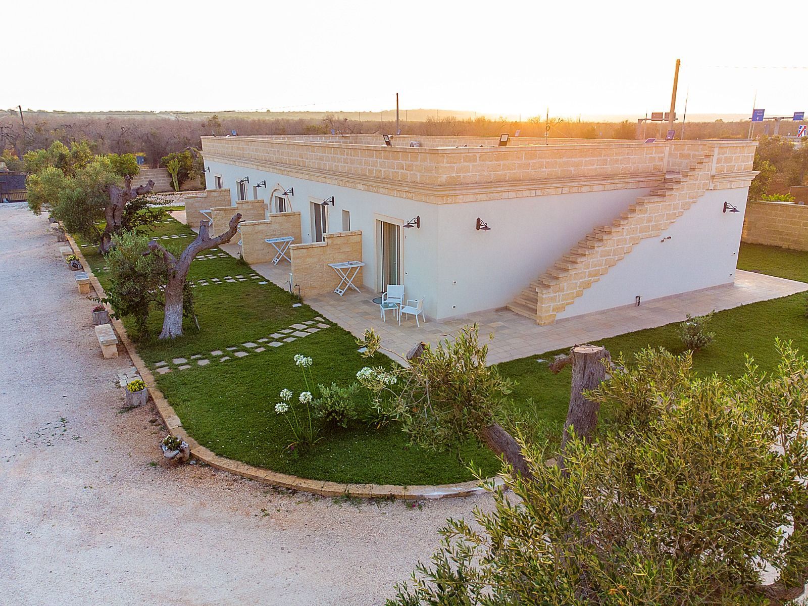 Edificio bianco con posti a sedere all'aperto, situato in una zona erbosa con sentieri in pietra e ulivi.