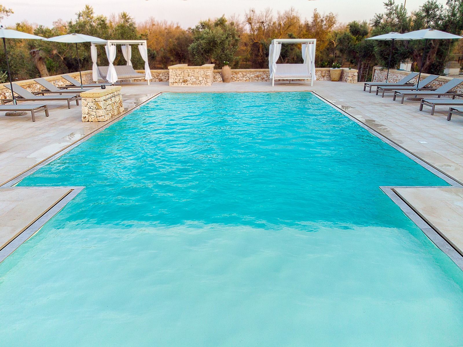 Piscina con acqua turchese, circondata da sedie a sdraio e cabine in un ambiente esterno soleggiato.