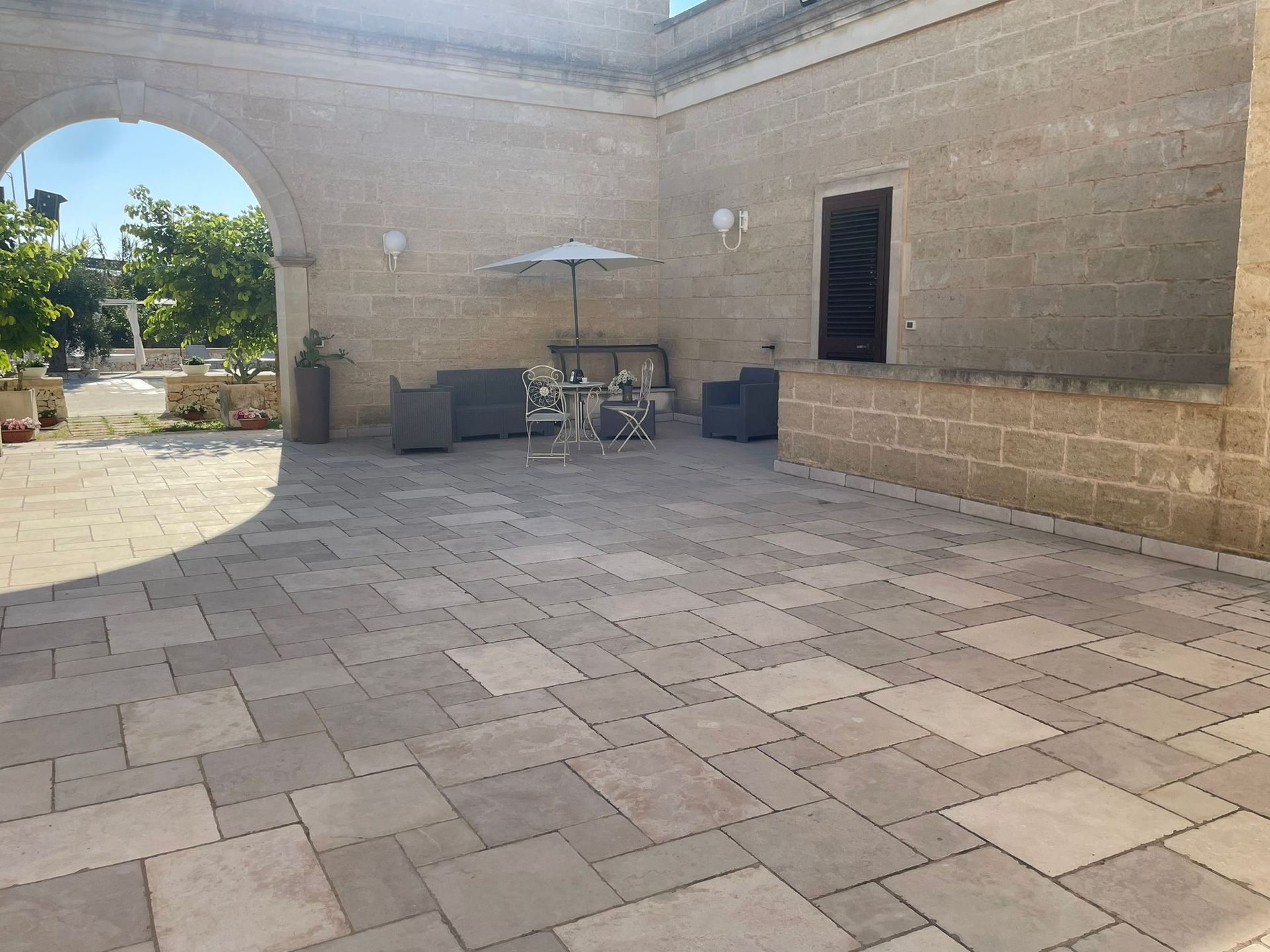 Patio in pietra con area salotto sotto un ombrellone, porta ad arco e pareti in pietra chiara in un cortile soleggiato.