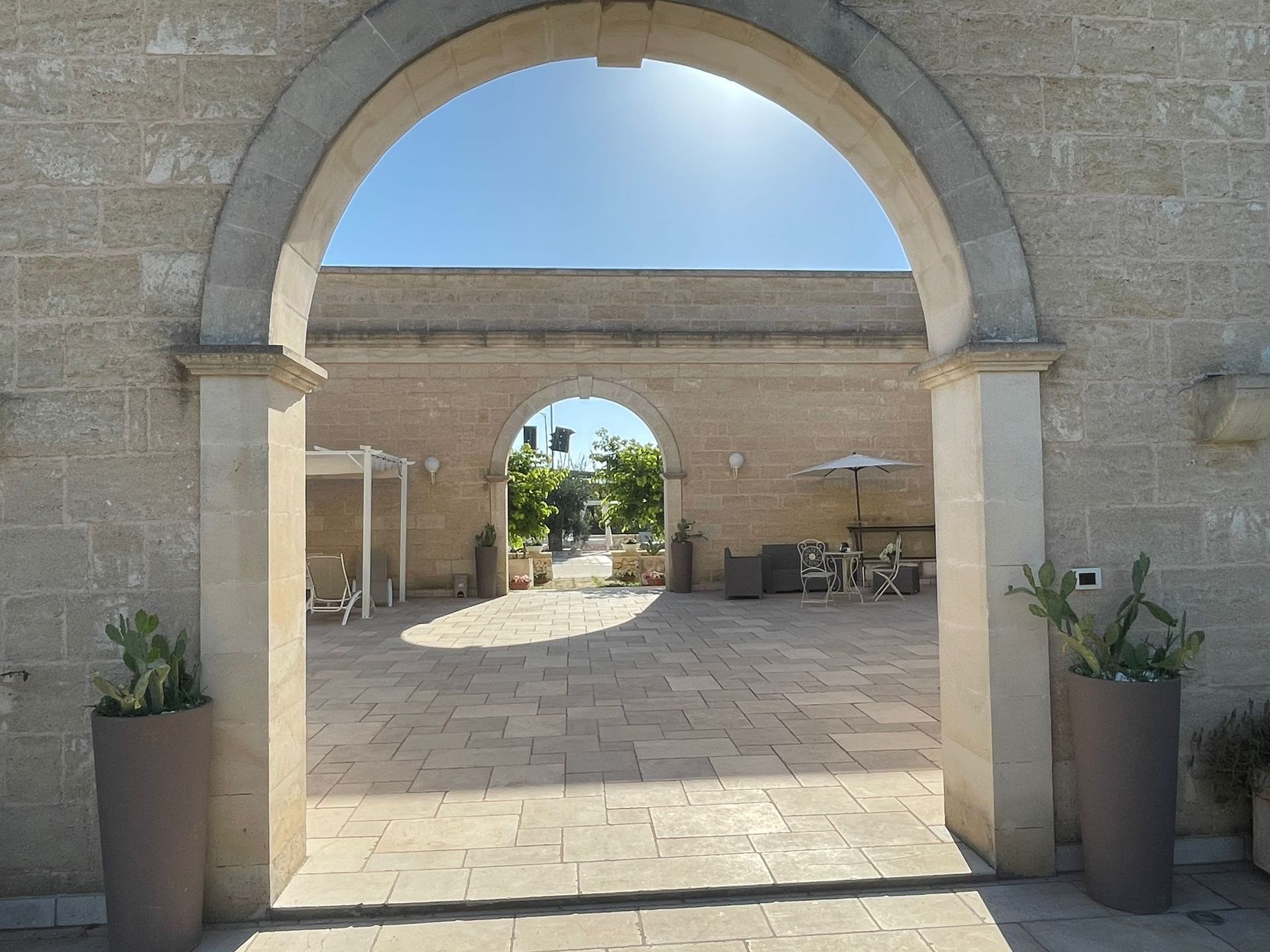 Un arco in pietra incornicia un cortile con tavoli, sedie, piante e un cielo azzurro e soleggiato.