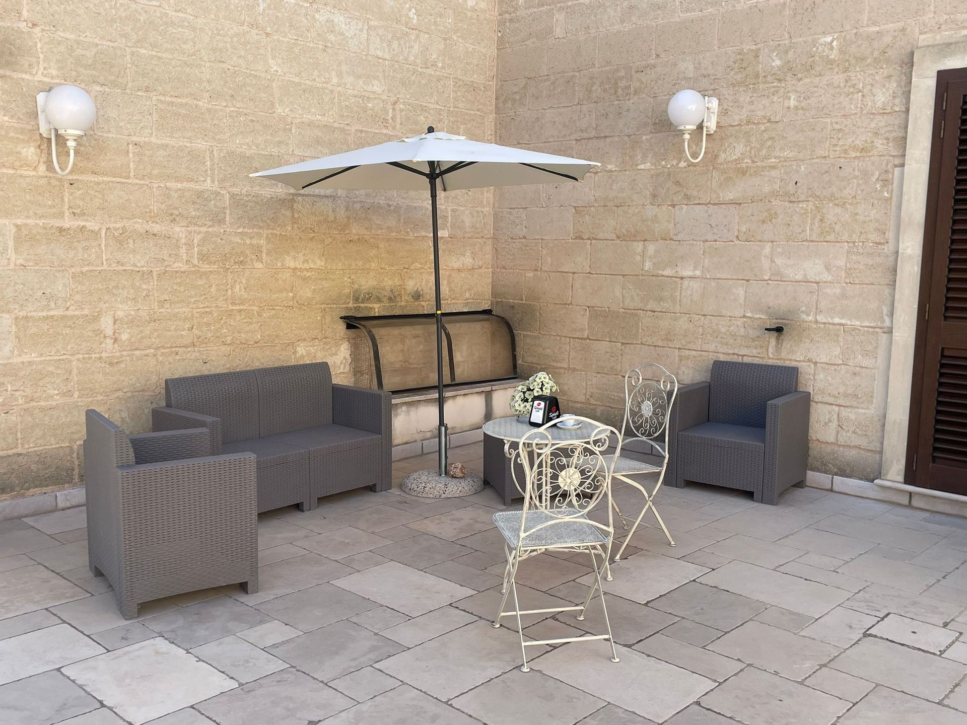 Area salotto all'aperto con mobili in vimini grigio, un tavolino e un ombrellone su un patio in pietra.