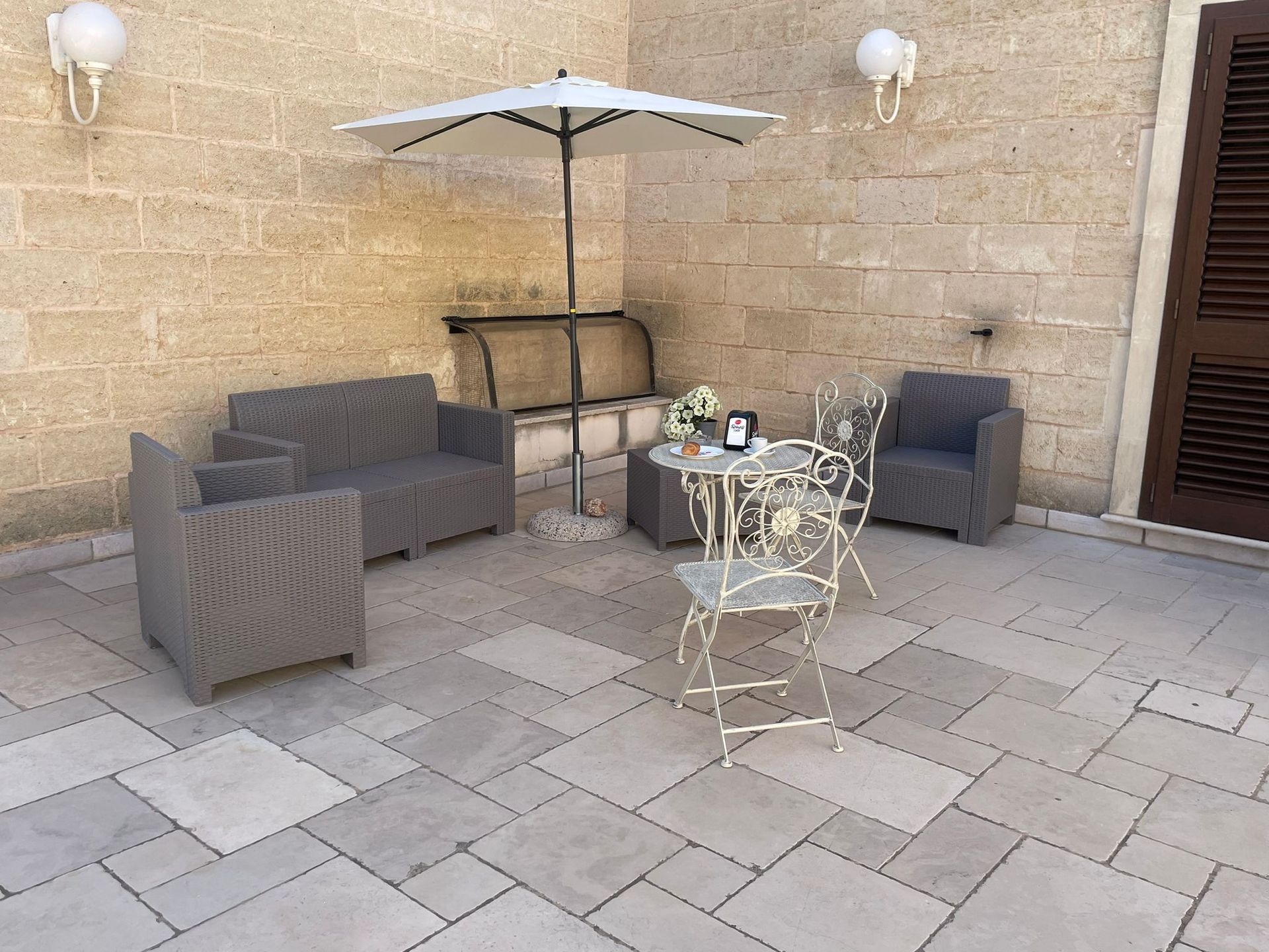 Patio con mobili grigi, un ombrellone e un tavolino contro un muro di pietra beige.