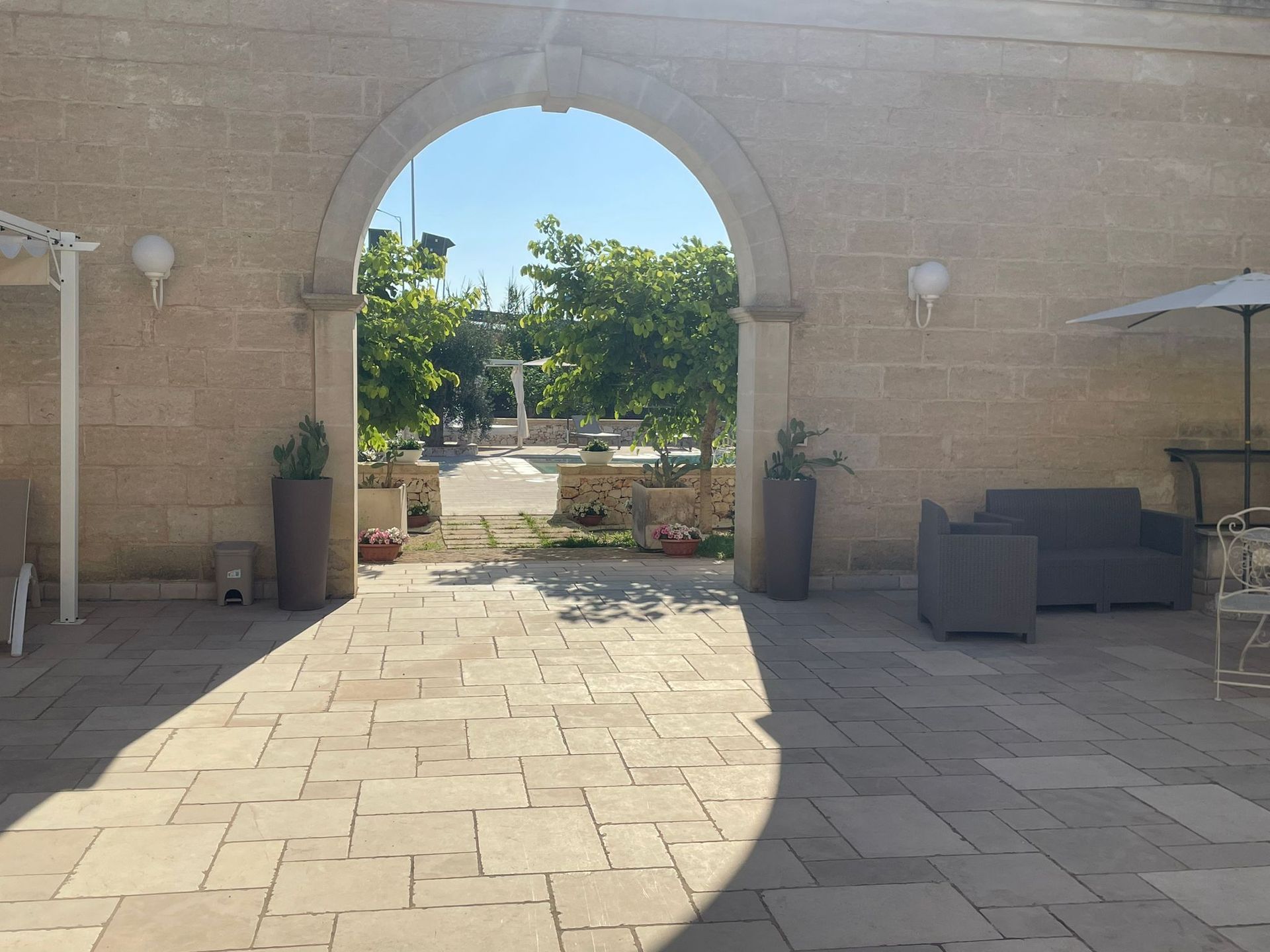 Un portale in pietra ad arco incornicia un patio soleggiato con alberi, mobili e piante in vaso.