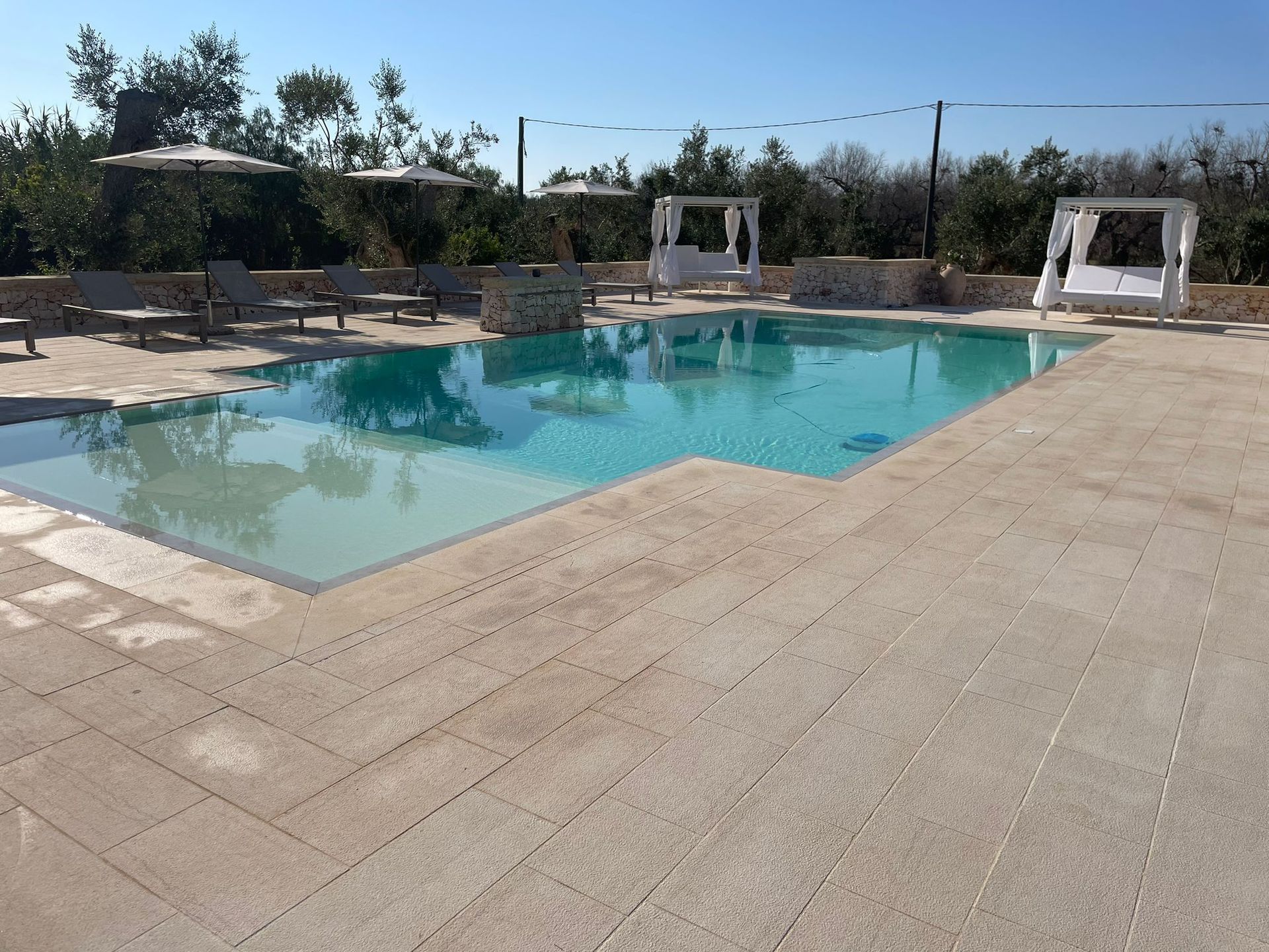 Piscina con sedie a sdraio bianche, cabine e patio in pietra beige sotto un cielo azzurro.