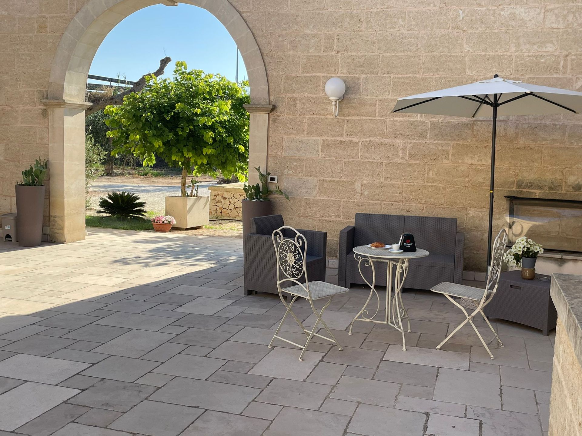 Patio in pietra con posti a sedere all'aperto sotto un ombrellone bianco e una porta ad arco che conduce a un giardino.