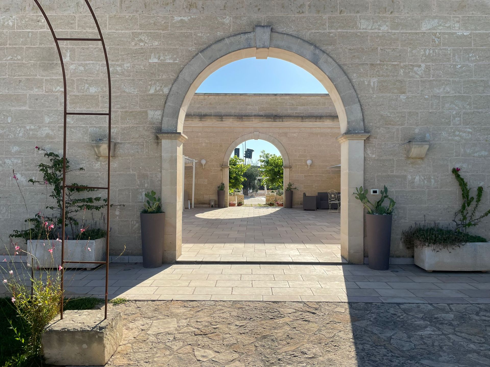Arco in pietra che si affaccia sul cortile, altri archi in lontananza, con piante in vaso e cielo soleggiato.