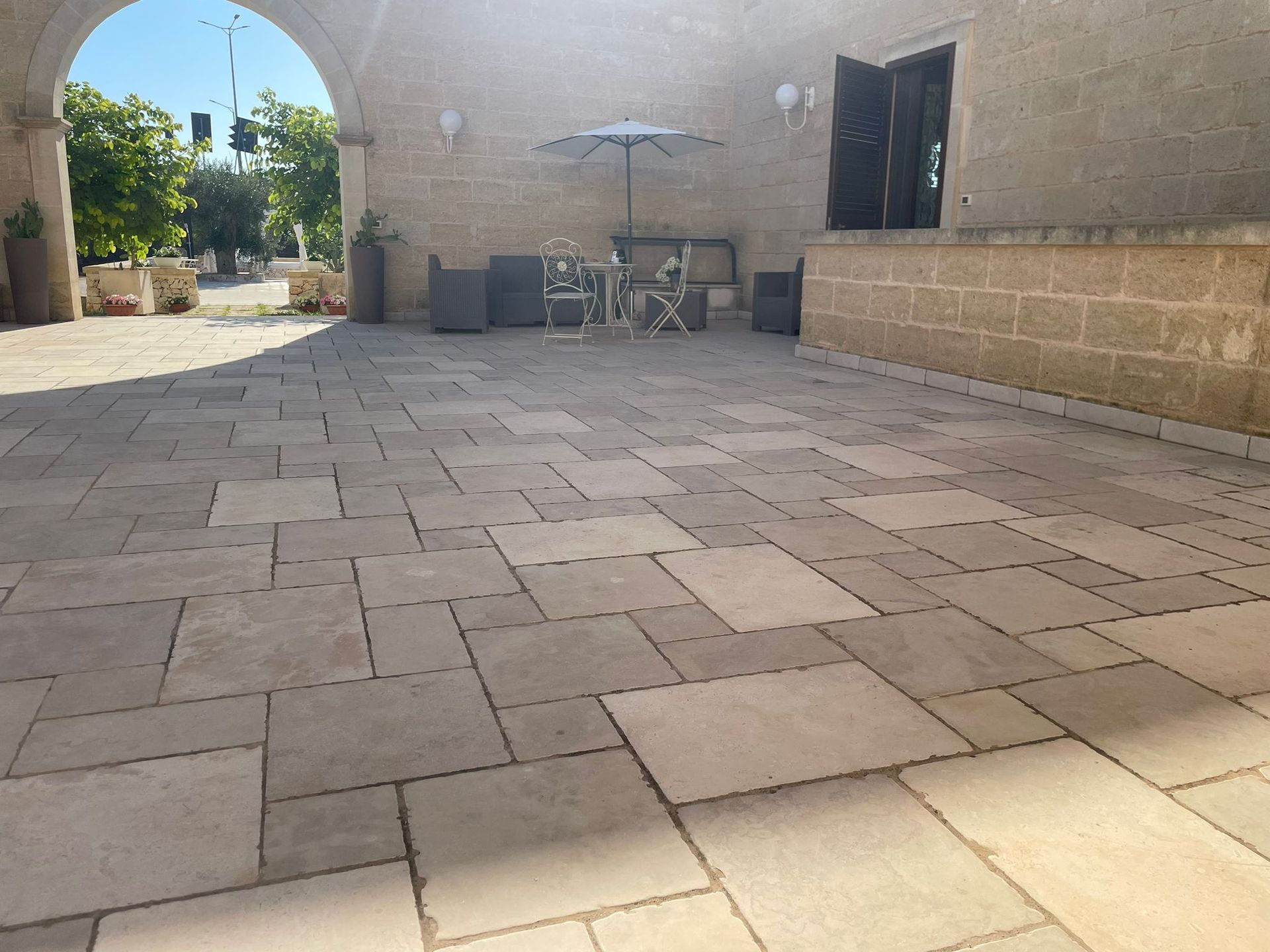 Patio lastricato in pietra con posti a sedere sotto un ombrellone. Sullo sfondo si vedono un arco in pietra e un edificio.