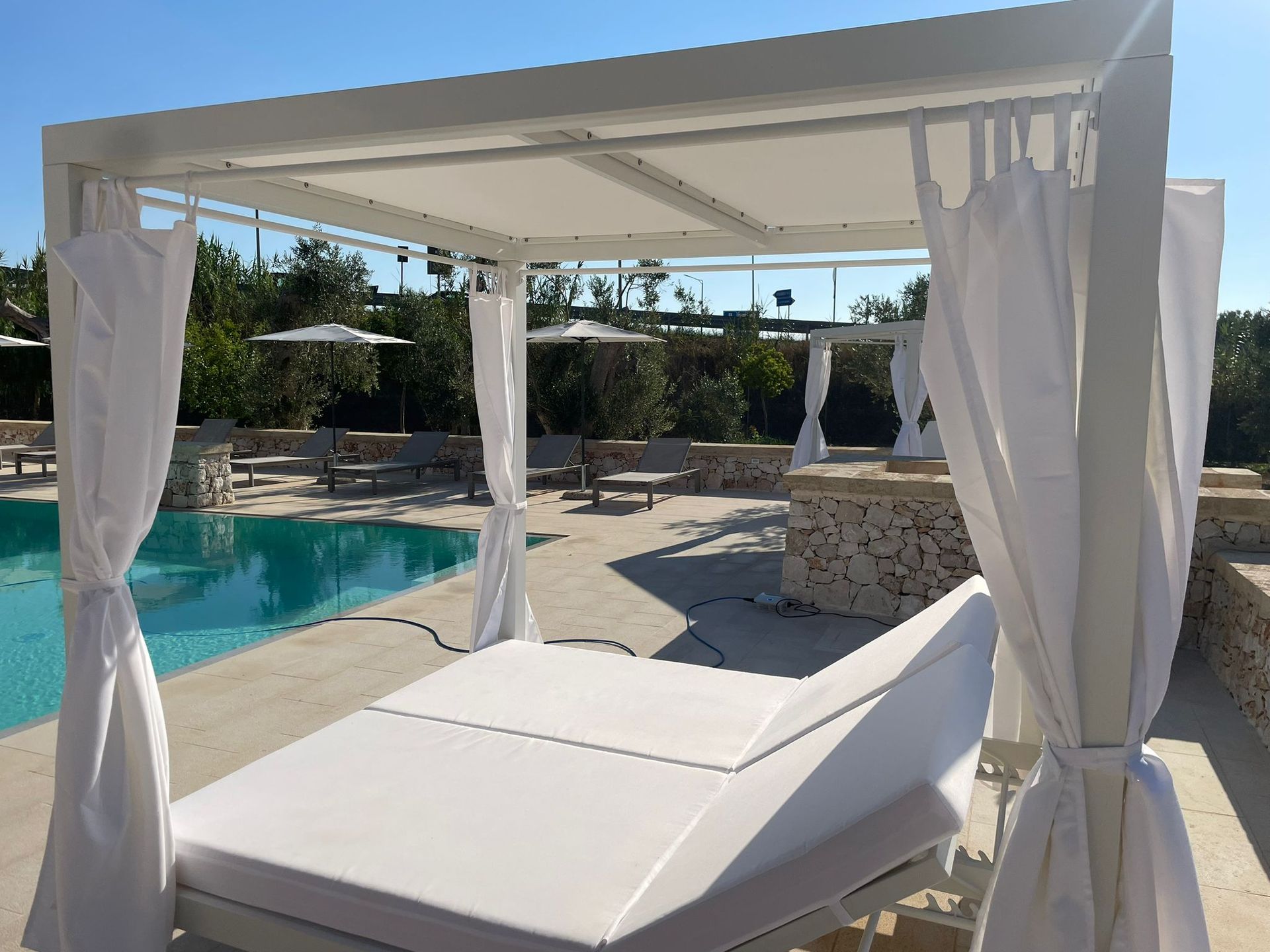 Letto bianco in stile cabana a bordo piscina con tende e ombrelloni.