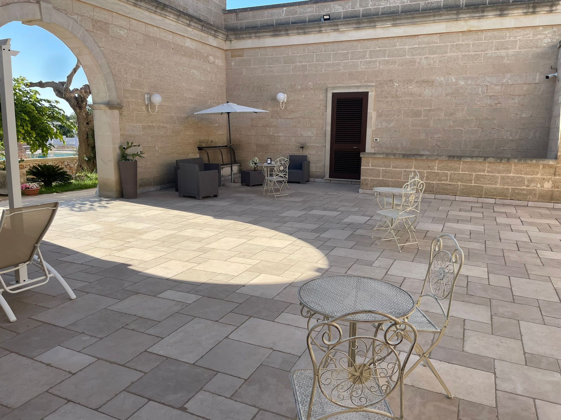 Patio esterno con pavimentazione in pietra, aree salotto e ombrellone parzialmente ombreggiato.