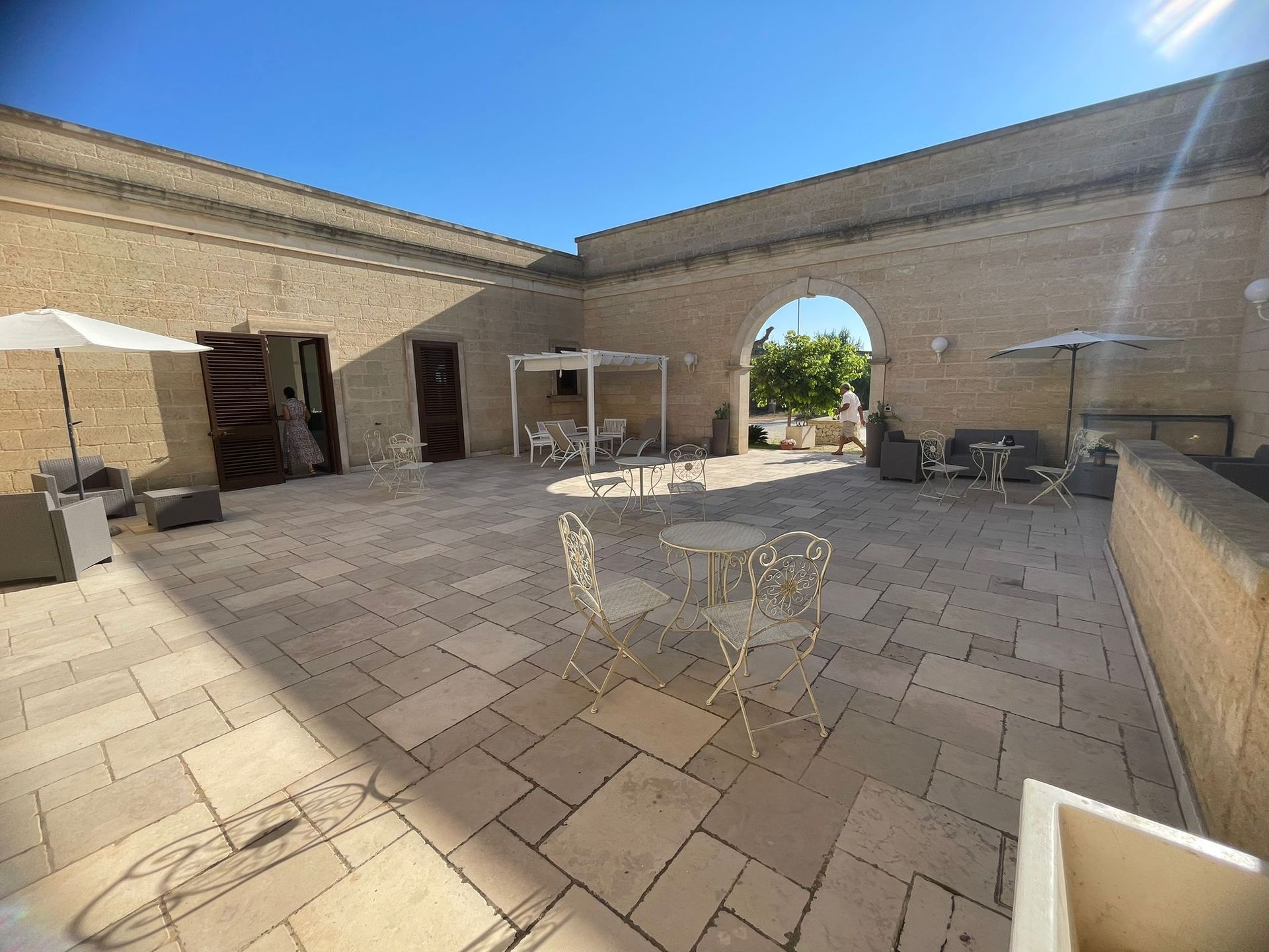Cortile con tavoli, sedie, ombrelloni e un arco, sotto un cielo azzurro brillante.