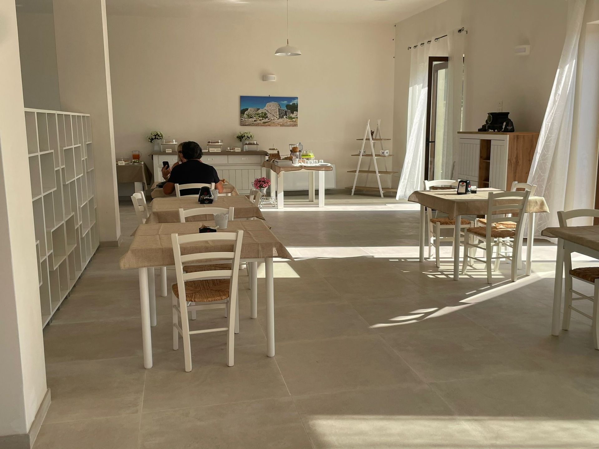 Ampia sala con tavoli, sedie e una persona seduta. Tonalità neutre e luminose, con luce naturale che filtra a fiotti.