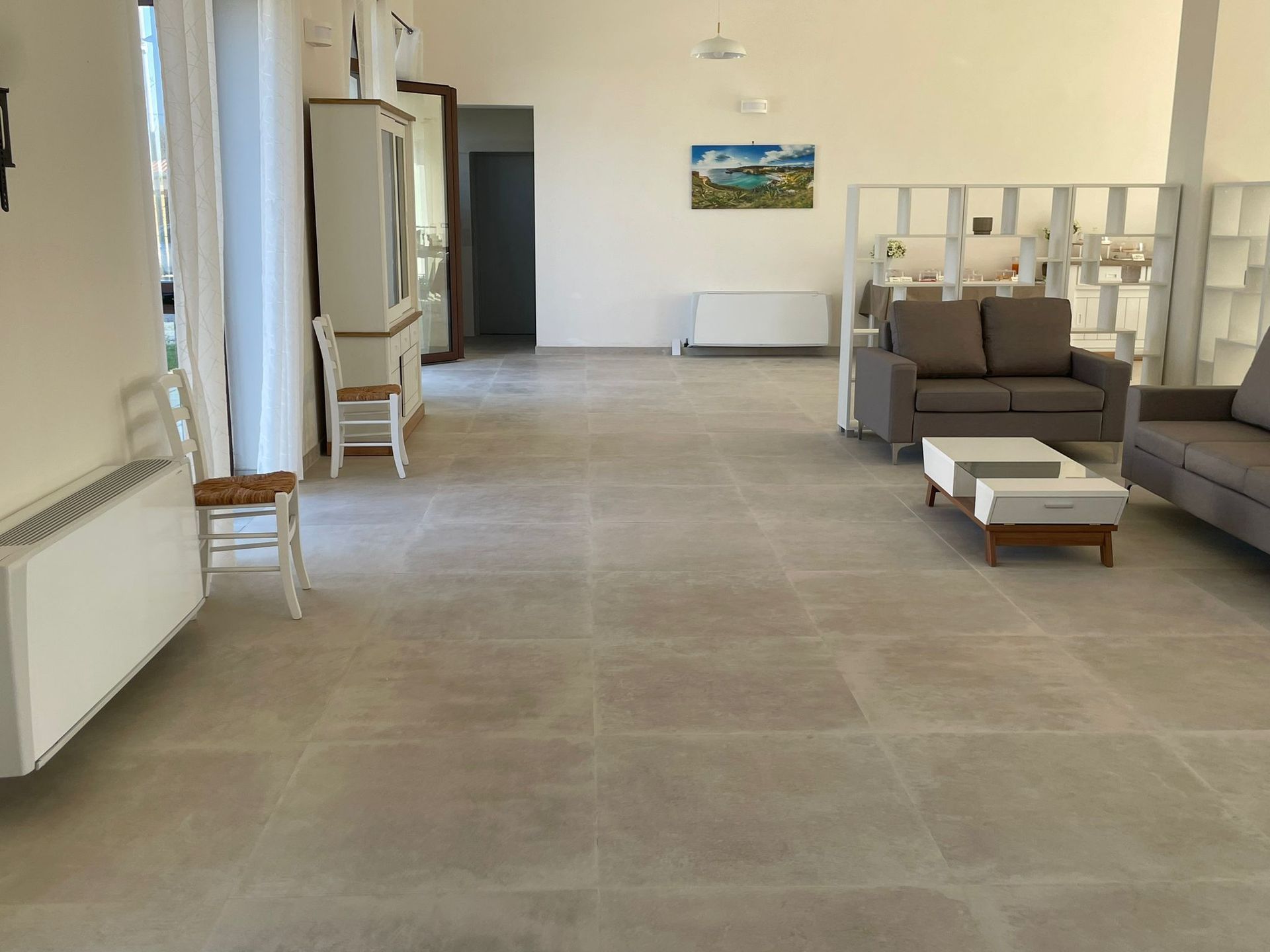 Ampio soggiorno open space con pavimento color pietra, divani e scaffalature bianche. Tonalità neutre, luminose e ariose.