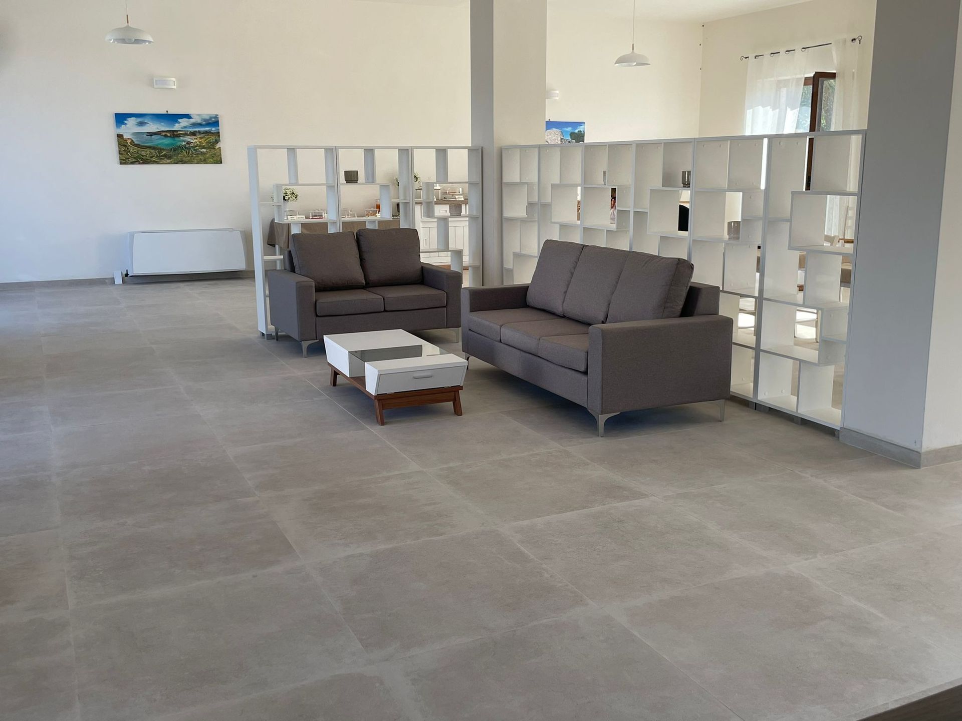 Zona soggiorno con divani grigi, un tavolino bianco e una parete divisoria decorativa. Pavimento piastrellato grigio.