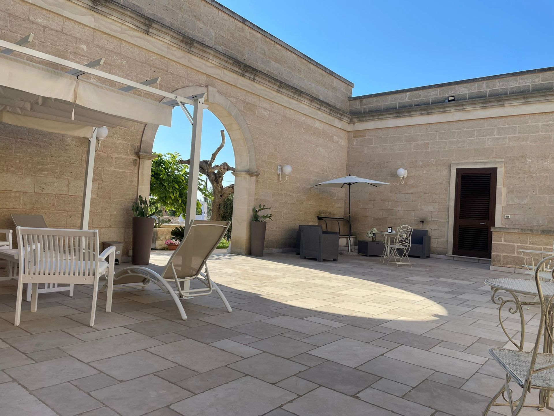 Patio esterno con muri in pietra, mobili bianchi e un arco aperto. Giornata di sole.