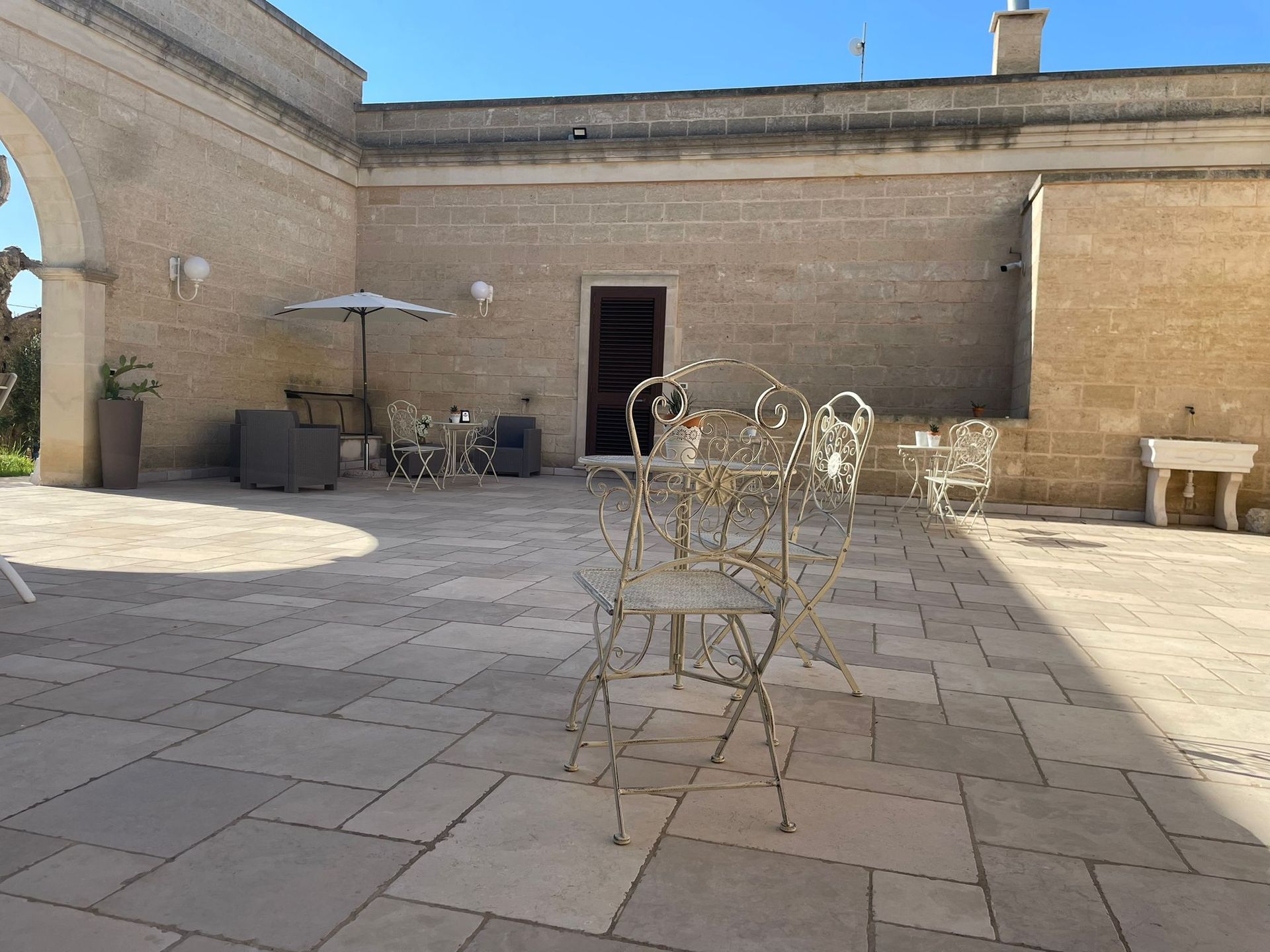 Patio con piastrelle in pietra, sedie, tavoli, ombrellone e muri in pietra beige.