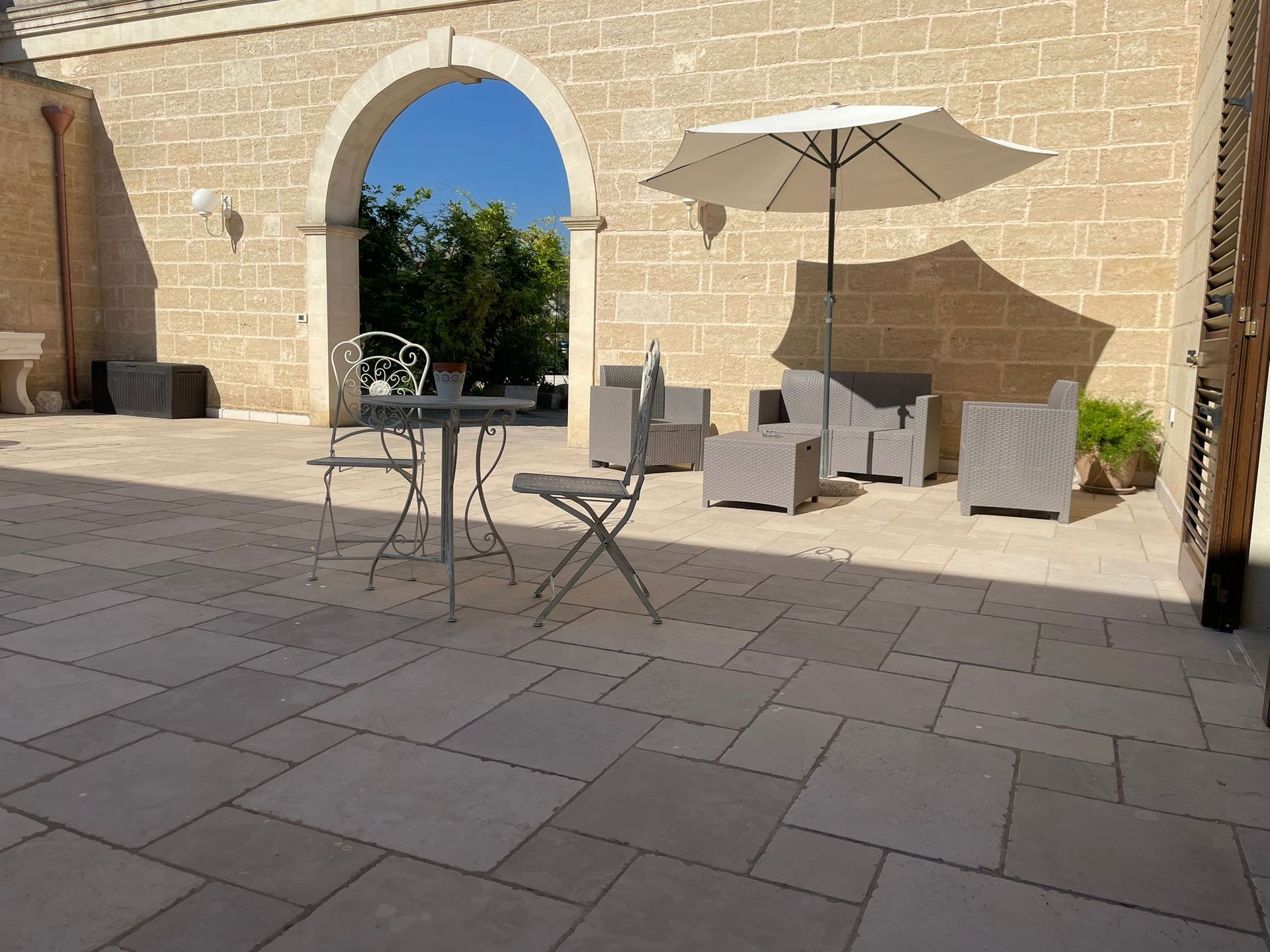 Patio con pavimento in pietra, posti a sedere e ombrellone. Porta ad arco che dà sull'esterno, soleggiato.