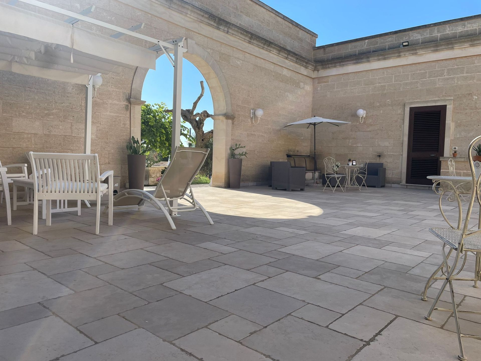 Cortile con pavimento in pietra, arco, tavoli e sedie bianchi e una poltrona reclinabile sotto una tenda.