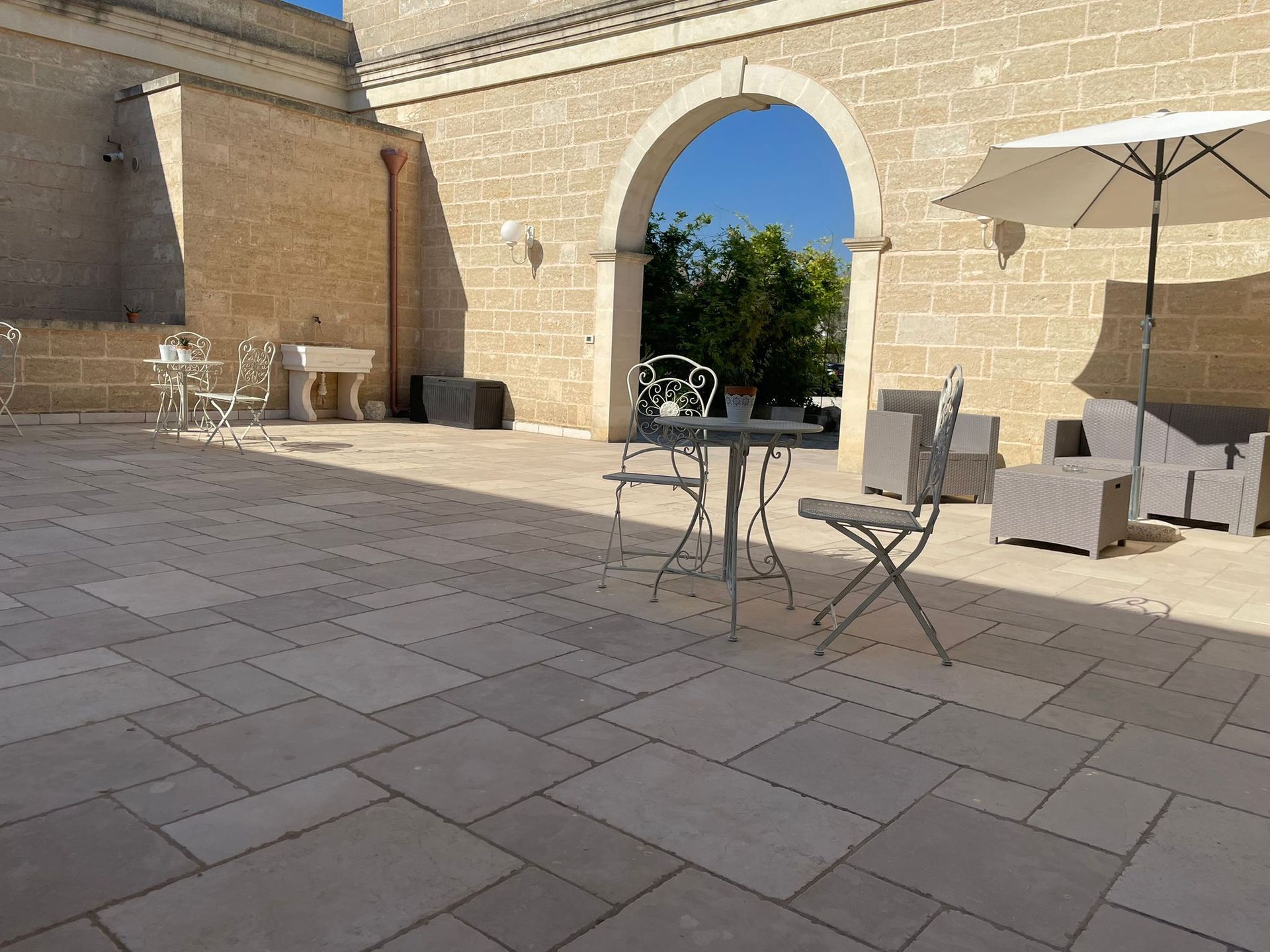Cortile con pavimentazione in pietra, posti a sedere e un portone ad arco che si affaccia su un cielo soleggiato.