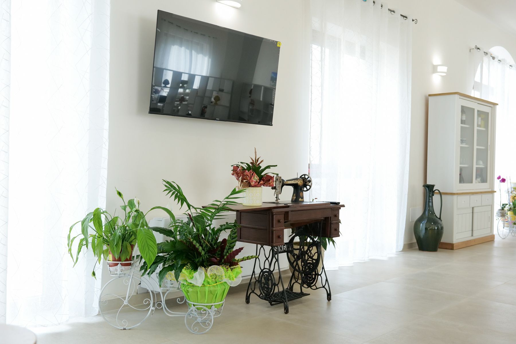 Stanza luminosa con macchina da cucire, piante, TV e mobiletto contro le pareti bianche.